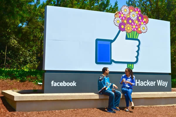 Couple sits under sign with a large "like button" and Facebook's logo